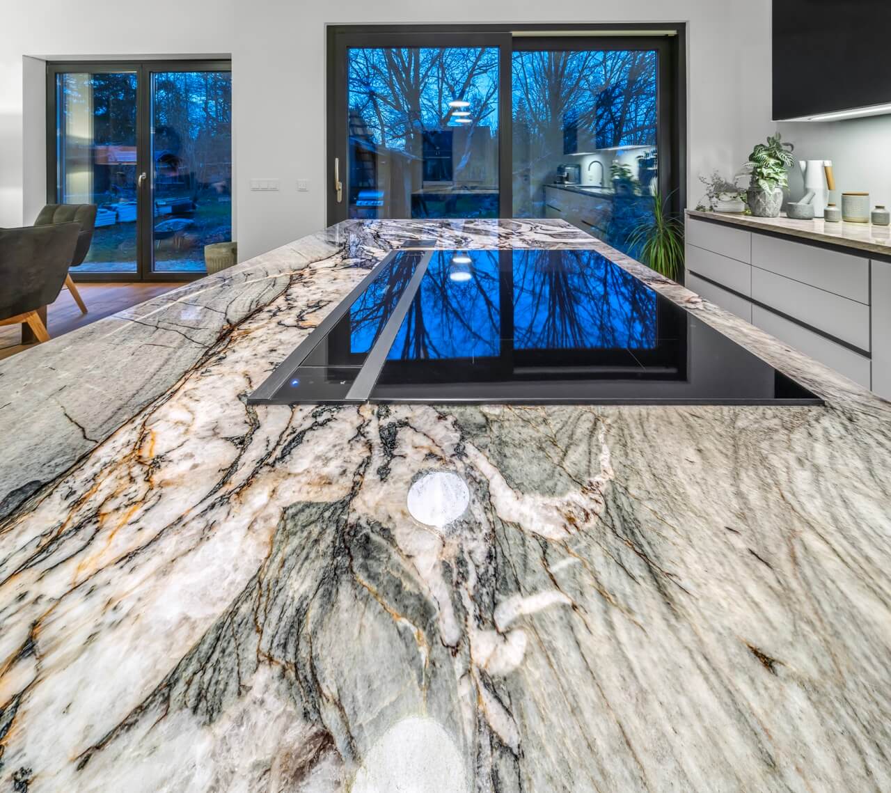 Naturstein Arbeitsplatten Berlin und Brandenburg: große Marmorinsel, Elektrokochfeld, Pflanzen, großes Fenster mit Ausblick.