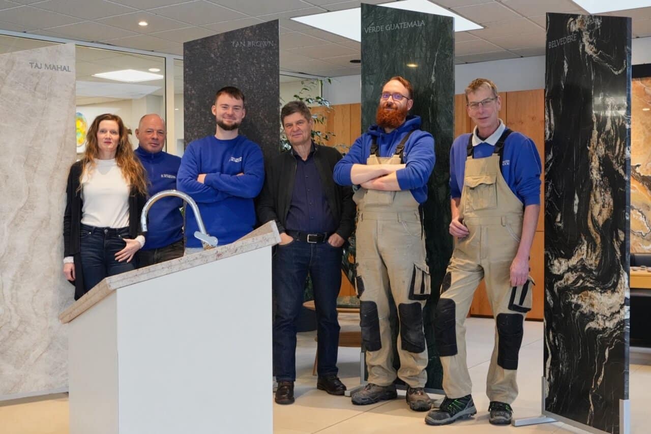 Sechs Personen vor großen Arbeitsplatten aus Naturstein Mustern mit Beschriftungen; vorne Display mit Wasserhahn, Innenaufnahme.