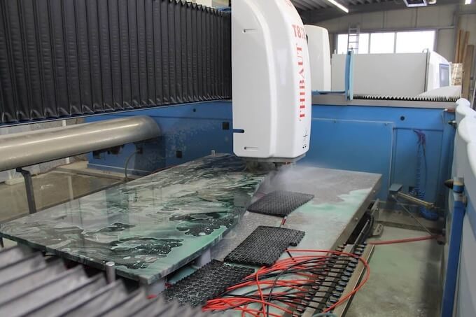 CNC Maschine schneidet Arbeitsplatten aus Naturstein in Berliner Werkstatt, Wasser und Schmutz, rote Kabel und schwarze Pads sichtbar.